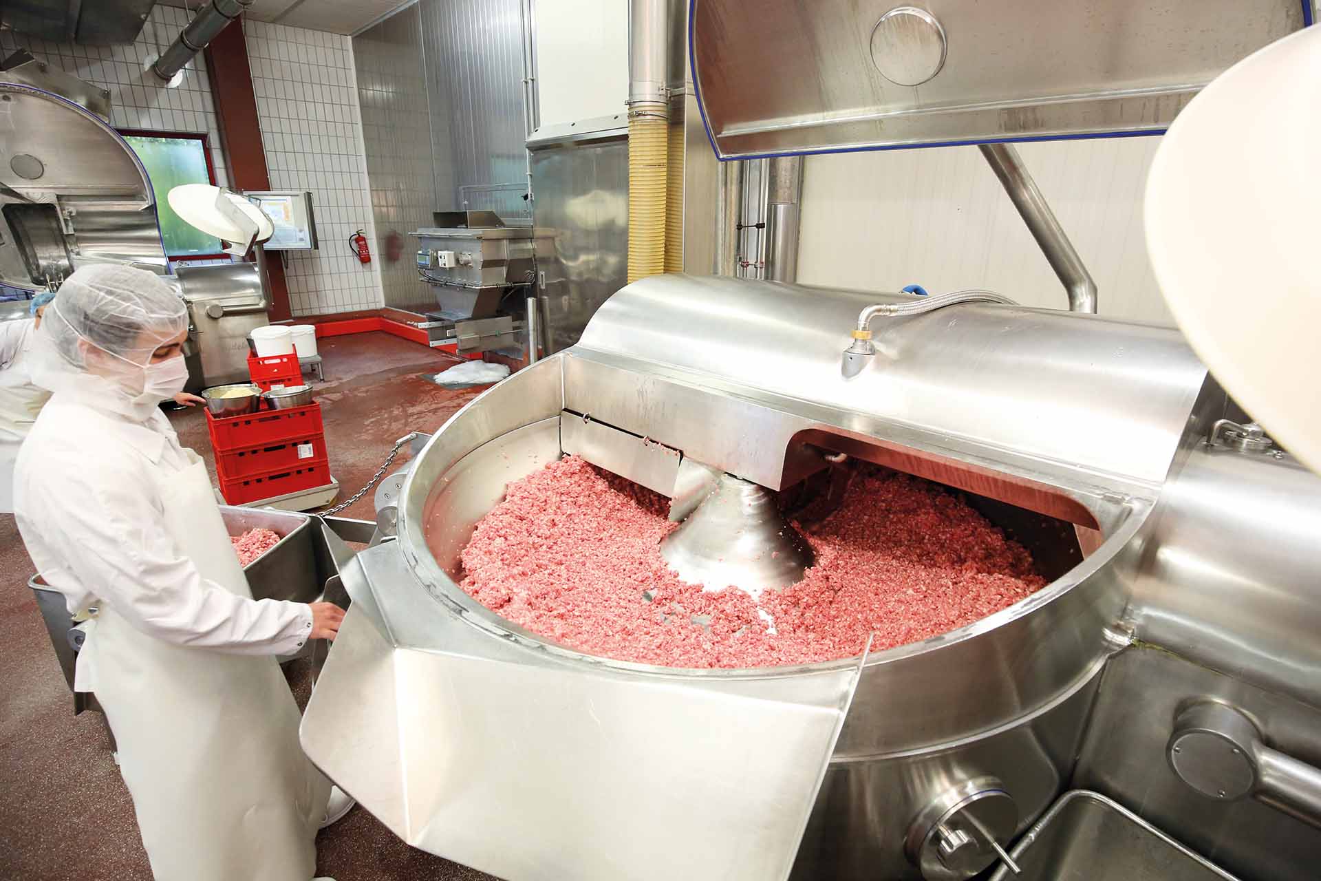 Person in Schutzkleidung neben großem Fleischmischer in Lebensmittelverarbeitungsbetrieb mit Edelstahlmaschinen und Fliesenwänden.